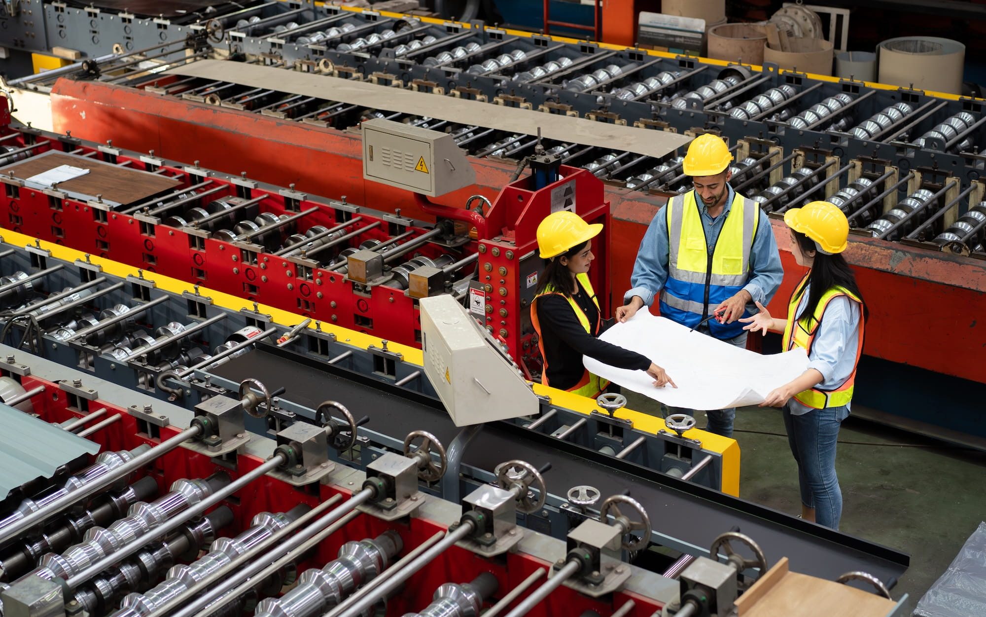 three people in safety gear stand beside a large metal forming machine reviewing a blueprint together in an active manufacturing environment