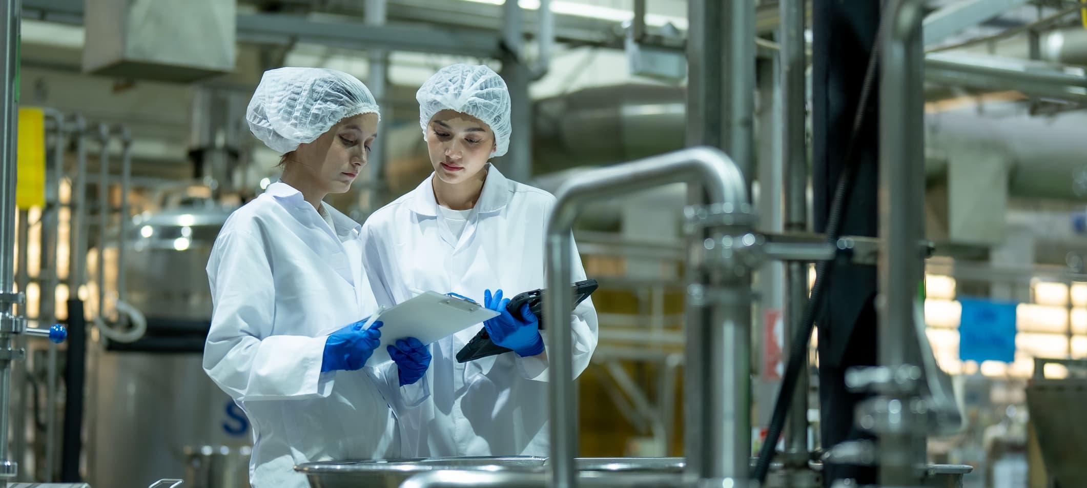 two technicians in protective gear verify ingredient accuracy for a beverage mixing process using a tablet and clipboard
